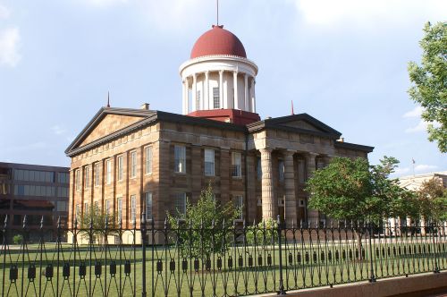 Historic building with a red dome, stone facade, and columns, surrounded by greenery and a decorative fence.