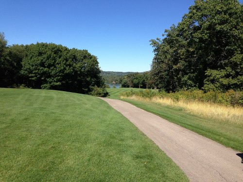 A scenic view of a golf course path lined with trees, leading towards a lake in the distance under a clear blue sky.