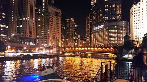 A nighttime cityscape featuring illuminated buildings along a river, with a boat cruising through the water.