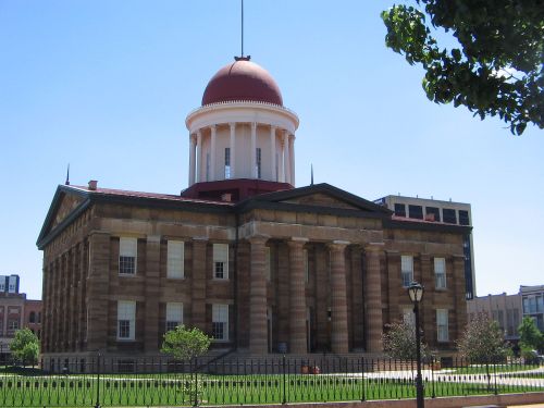 Historic building with a red dome, columns, and a clear blue sky, surrounded by green trees and a fence.