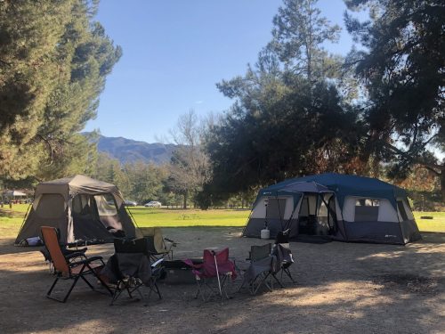 A campsite with two tents and several folding chairs set up under trees, with mountains in the background.
