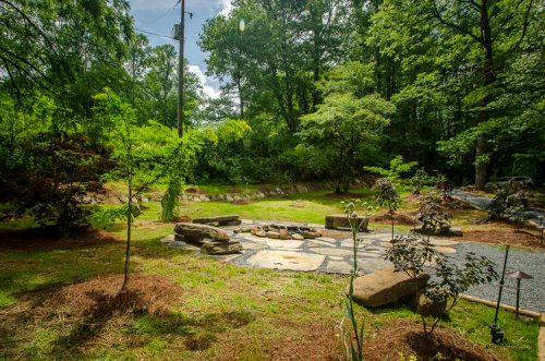 A serene garden with stone pathways, surrounded by trees and greenery, featuring a small fire pit area.