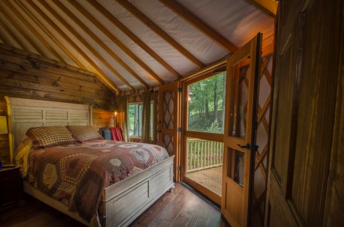Cozy bedroom with a wooden bed, quilted bedding, and large windows opening to a forest view. Warm, rustic decor.