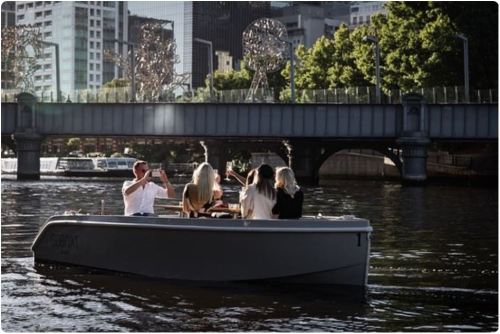 A group of four people on a boat enjoying drinks, with a city skyline and bridge in the background.