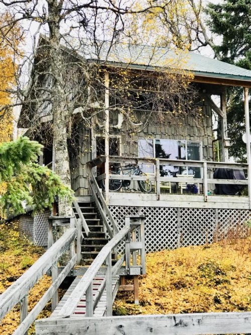 A rustic wooden cabin surrounded by autumn foliage, featuring a porch and stairs leading up to the entrance.