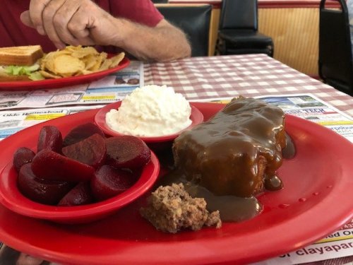 A red plate with meatloaf covered in gravy, mashed potatoes, beets, and a side of bread on a checkered tablecloth.
