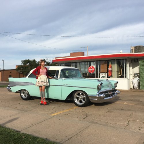 A woman in a red cardigan and skirt stands next to a vintage mint green car in front of a retro diner.
