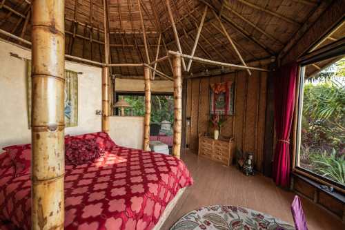 Cozy bamboo interior of a tropical bedroom with a red patterned bedspread and large windows overlooking greenery.