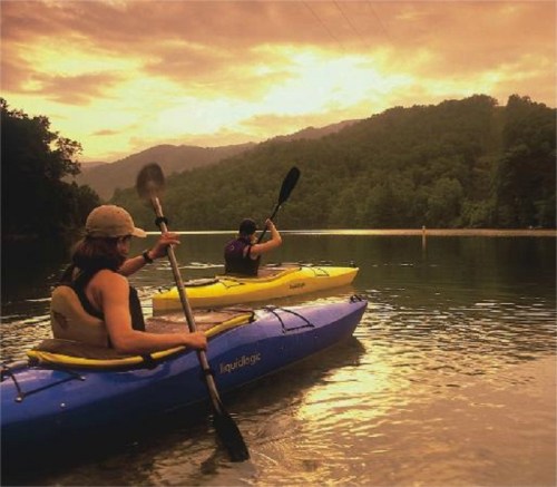 Two people kayaking on a calm lake at sunset, surrounded by mountains and trees.