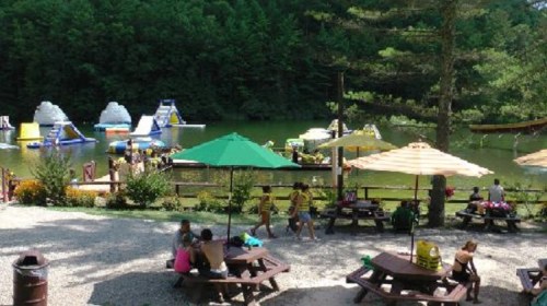 A lakeside scene with inflatable water attractions, picnic tables, and umbrellas, surrounded by trees and people enjoying the day.