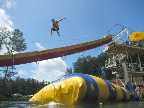 A child in a life jacket jumps off a platform into a large inflatable water blob at a lake.