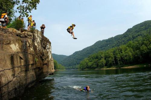 A child jumps off a rocky ledge into a river while others watch from above, surrounded by lush green hills.