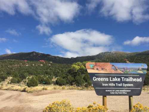 Sign for Greens Lake Trailhead, surrounded by mountains and greenery under a partly cloudy sky.