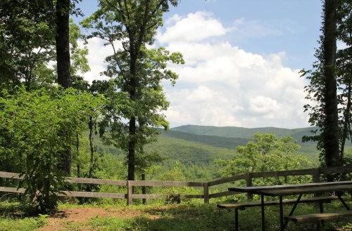 A scenic view of rolling green hills and trees under a partly cloudy sky, with a wooden fence and picnic table in the foreground.