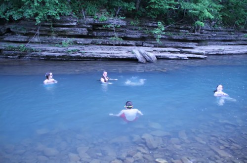 Four people swimming in a clear blue river surrounded by rocky banks and lush greenery.