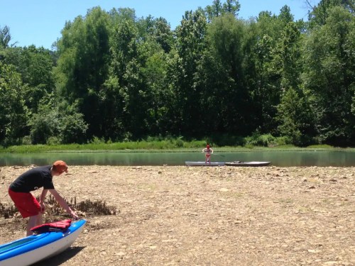 A sunny riverside scene with two people near kayaks, surrounded by lush green trees and a calm waterway.