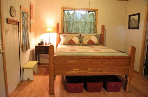 Cozy bedroom with a wooden bed, quilted bedding, nightstand, lamp, and storage bins under the bed. Natural light from window.