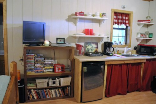 Cozy kitchen with a TV, shelves of dishes, coffee maker, microwave, and a small fridge, featuring red accents.