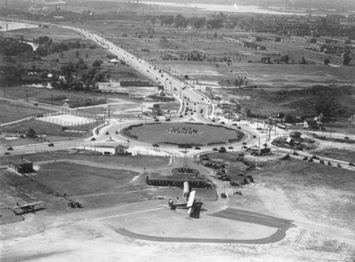 Aerial view of a roundabout with cars, an airplane on the ground, and surrounding fields and roads.
