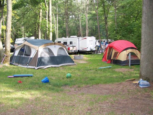 Two tents set up in a grassy campsite surrounded by trees, with a camper in the background and scattered toys on the ground.
