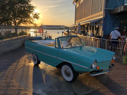 A vintage aqua convertible car parked by the water at sunset, with a restaurant and an American flag in the background.