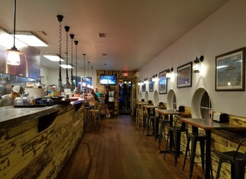 Interior of a cozy restaurant with wooden floors, bar seating, and framed pictures on the walls. Chefs are visible in the kitchen.
