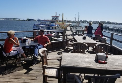Two people sit at a table on a waterfront deck, with boats and a bustling pier in the background under a clear blue sky.