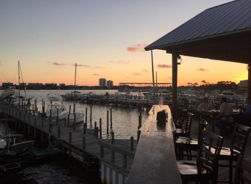 Sunset over a marina with boats docked, viewed from a wooden deck with chairs and tables.