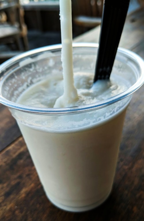A clear plastic cup filled with a creamy beverage, featuring a black straw and spoon, set on a wooden table.