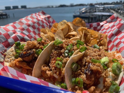 Three tacos topped with green onions and crispy bits, served with potato chips, against a scenic waterfront backdrop.