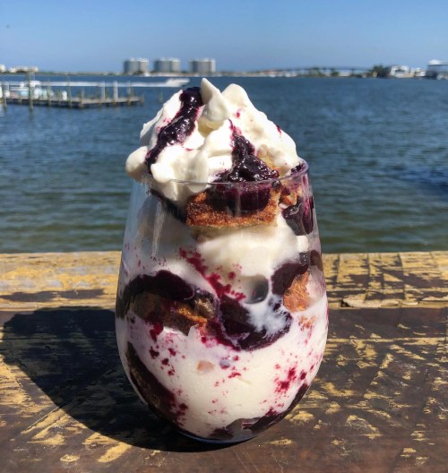 A tall glass filled with layers of ice cream, cake, and berry sauce, topped with whipped cream, against a scenic waterfront backdrop.