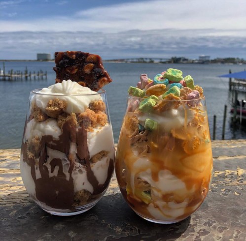 Two dessert glasses filled with layers of chocolate and vanilla ice cream, topped with whipped cream and colorful cereal, by a waterfront.