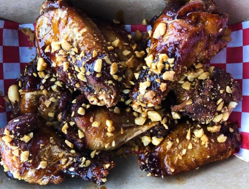 A box of glazed chicken wings topped with crushed peanuts on a red and white checkered paper.