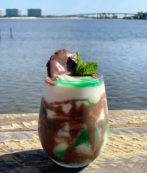 A layered dessert in a glass with chocolate, mint, and cream, set against a scenic waterfront view.