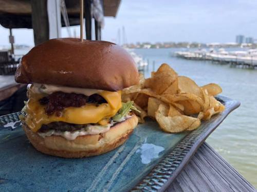 A juicy cheeseburger with lettuce and sauce, served with potato chips, against a scenic waterfront backdrop.