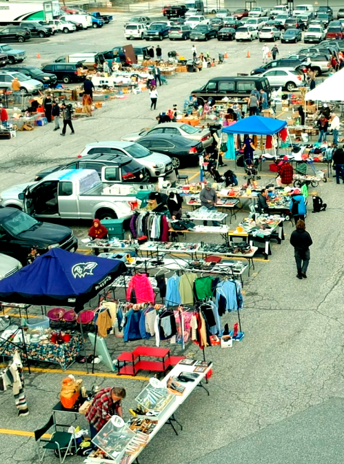 Aerial view of a bustling flea market with various stalls, cars parked nearby, and people browsing items.