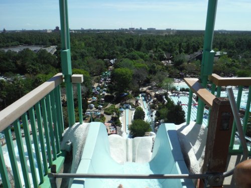 View from the top of a water slide, overlooking a lush landscape and a busy water park below.