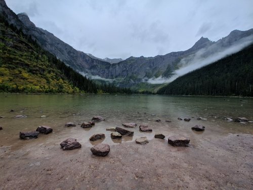 A serene lake surrounded by mountains, with mist and rocky shoreline under a cloudy sky.