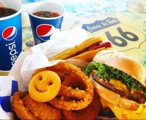 A colorful meal featuring a burger, hot dogs, onion rings, and two cups of soda on a Route 66-themed table.