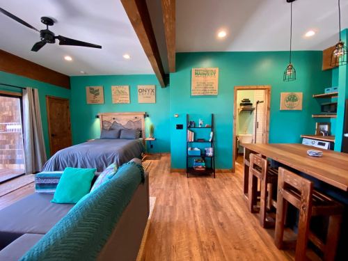 A vibrant teal room featuring a bed, seating area, wooden accents, and a small kitchenette with a bathroom visible.