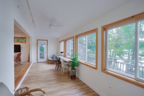 Bright, airy room with large windows, wooden floors, and a view of greenery outside. Cozy seating and a small table included.