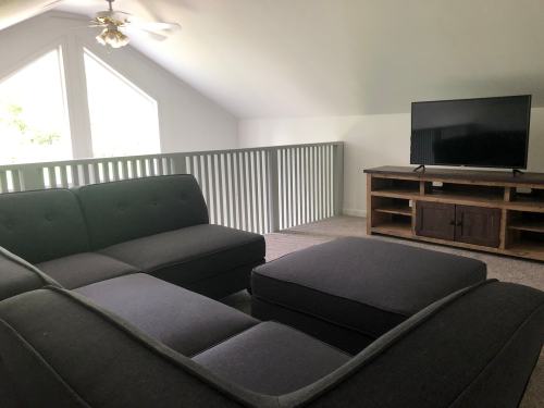 Cozy loft space with a gray sectional sofa, ottoman, and a TV on a wooden stand, featuring large windows for natural light.