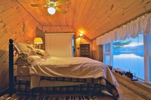 Cozy attic bedroom with wooden walls, a bed, and a window overlooking a serene view, illuminated by warm lighting.