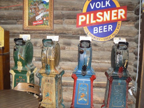 Four vintage slot machines in a rustic setting, with a sign for Volk Pilsner Beer in the background.
