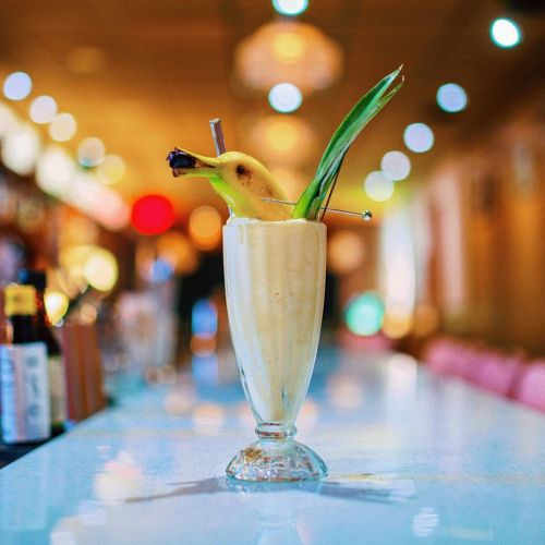 A tropical cocktail in a tall glass, garnished with a banana and pineapple leaf, set against a blurred bar background.