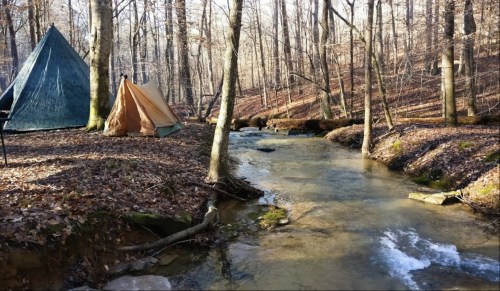 Two tents by a clear stream in a wooded area, surrounded by trees and fallen leaves in early spring.