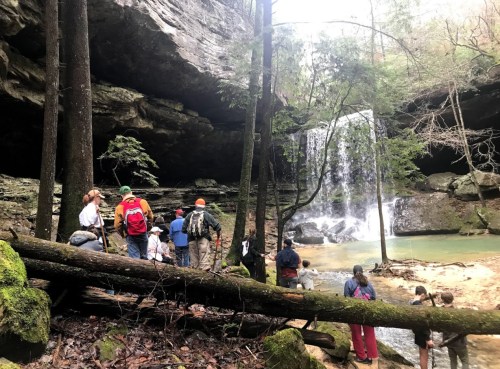A group of people gathered near a waterfall in a forested area, surrounded by rocks and trees.
