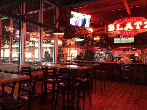 A vibrant bar interior with red neon signs, wooden tables, and TVs showing sports, creating a lively atmosphere.