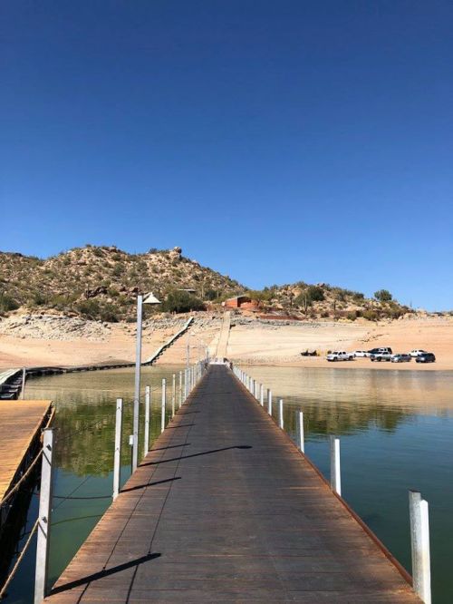 A wooden dock extends into calm water, leading to a sandy shore and hills under a clear blue sky.