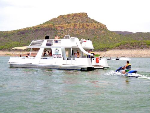 A houseboat on a lake with people onboard, while a person rides a jet ski nearby against a hilly backdrop.